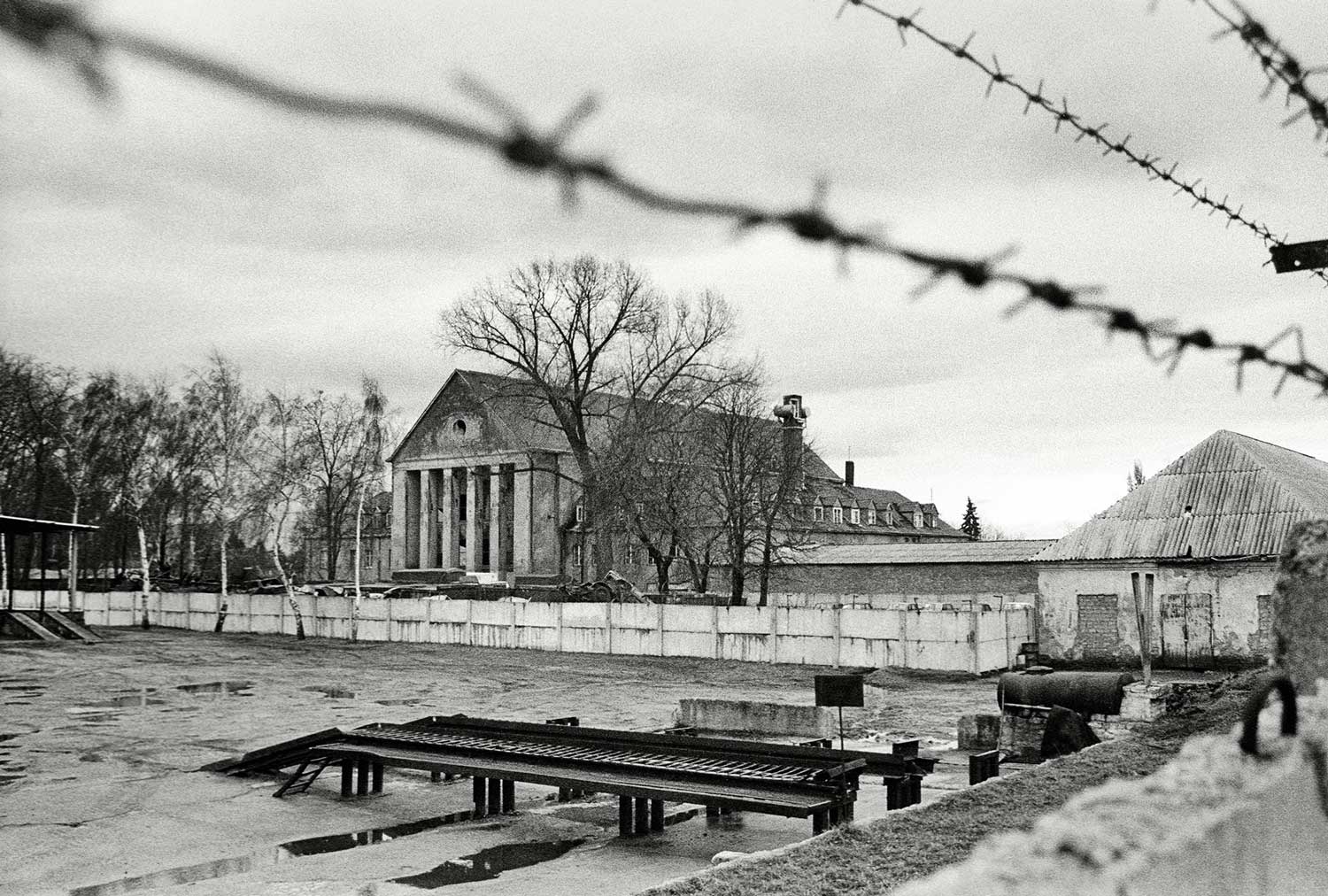 Foto, schwarz-weiß: Blick über eine Mauer mit Stacheldraht auf das Festspielhaus im Hintergrund, davor ein Hof mit flachen Bänken.