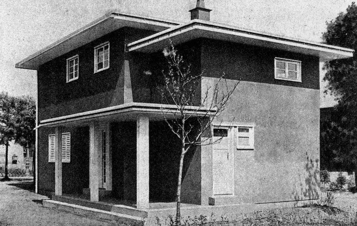 Foto, schwarz-weiß: Ein zweigeschossiger Bungalow mit Flachdach und Veranda