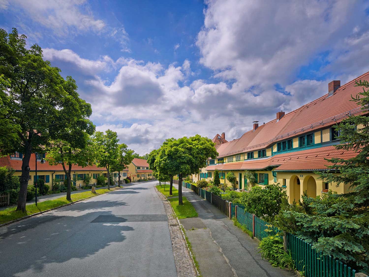 Foto, farbig, außen: Straßenzug mit gelb gestrichenen, zweigeschossigen Häusern mit Giebeldach hinter belaubten Straßenbäumen unter bewölktem Himmel.