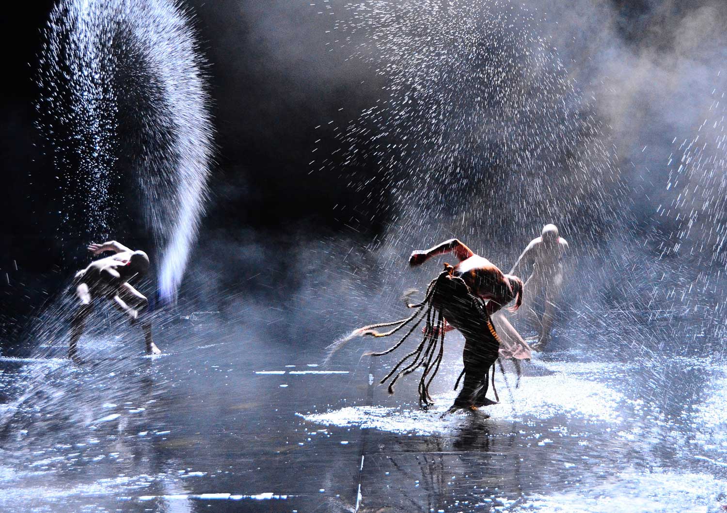Foto, farbig, innen: Drei Personen stehen auf dem knöcheltief mit Wasser gefüllten Boden und schlagen mit wilden Bewegungen Wasserfontänen heraus.
