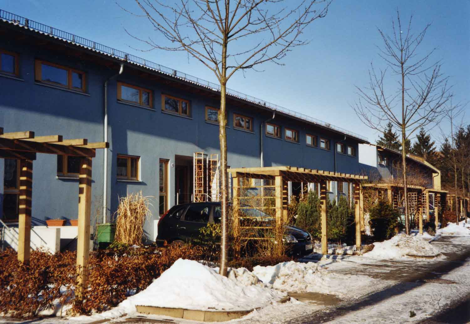Foto, farbig: Drei Reihenhäuser mit Vorgärten, kahlen Straßenbäumen und Schnee auf dem Gehweg unter blauem Himmel.