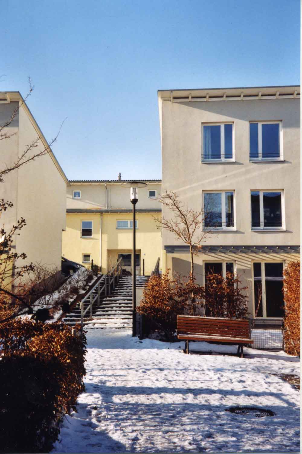 Foto, farbig: Rechts ein kleines dreigeschossiges Haus mit Flachdach, links eine Freitreppe, davor braun belaubte Büsche und eine Parkbank, es liegt Schnee, der Himmel ist wolkenlos.