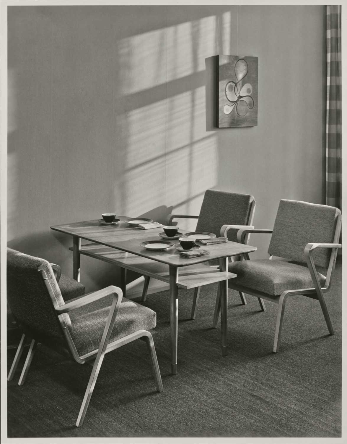 Foto, schwarz-weiß: Vier gepolsterte Armlehnsessel stehen um einen gedeckten, rechteckigen Tisch.